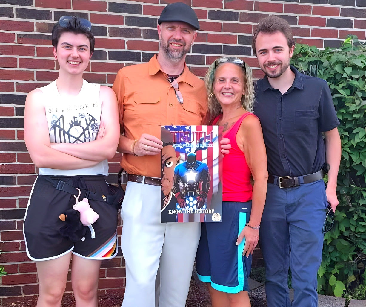 Family of four standing in front of a brick wall holding a promo poster for the comic project 'TRUTH: Know the History' with an American flag background design.